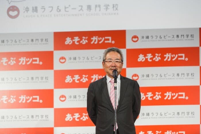 Nobuhiko Horie sale sul palco di una conferenza stampa annunciando il progetto di “Okinawa Rough & Peace College”